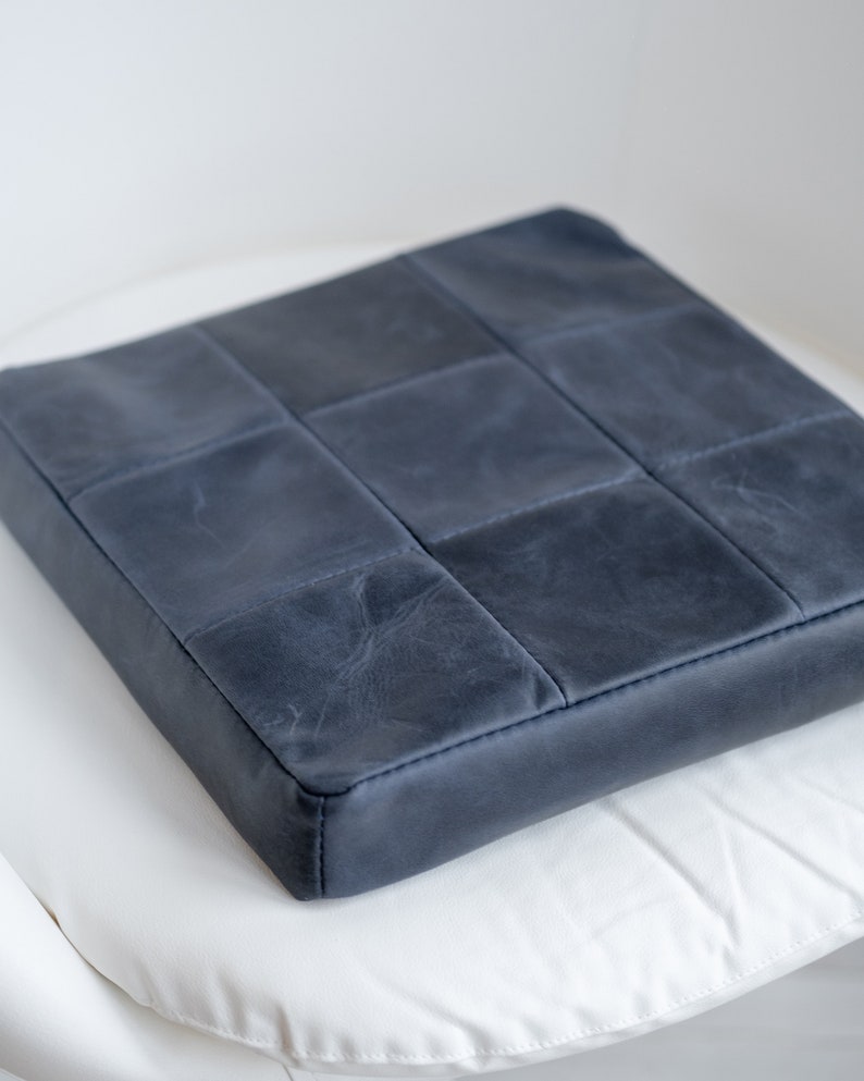May include: A square, navy blue cushion with a quilted design. The cushion is made of a leather-like material and has a stitched border. The cushion is placed on a white surface.