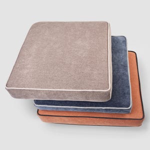 May include: A stack of three square cushions in neutral colors. The top cushion is a light brown with a white trim. The middle cushion is a dark blue with a white trim. The bottom cushion is orange with a black trim. The cushions are likely for furniture.