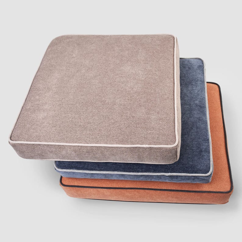 May include: A stack of three square cushions in neutral colors. The top cushion is a light brown with a white trim. The middle cushion is a dark blue with a white trim. The bottom cushion is orange with a black trim. The cushions are likely for furniture.