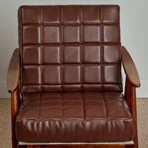 Op de afbeelding: Een bruine leren fauteuil met een gestoffeerde rugleuning en zitting. De stoel heeft een houten frame en armleuningen.