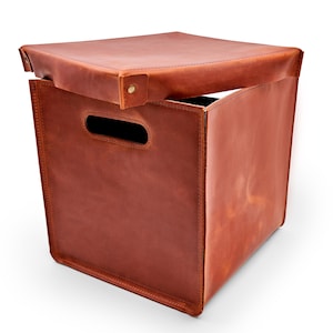 May include: A brown leather storage cube with a hinged lid. The cube features a rectangular handle cutout on one side. The lid is partially open, revealing the interior. The leather has visible stitching and a natural grain.