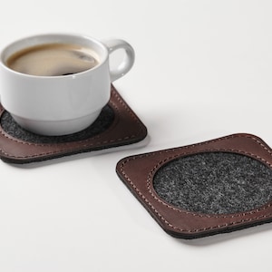 以下が含まれることがあります： グレーのフェルトインサートが付いた2つの茶色の革製コースター。1つのコースターには、コーヒーが入った白いコーヒーカップが置かれています。