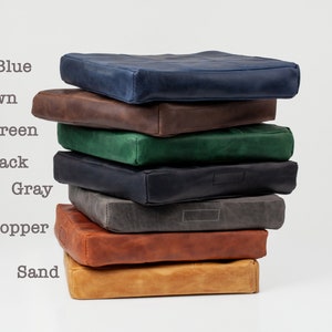 May include: A stack of square leather cushions in various colors: blue, brown, green, black, gray, copper, and sand. The cushions have a stitched design and are stacked on a white background. The word "Blue" is written above the blue cushion.
