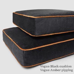May include: Two black square cushions with amber-colored piping. The cushions are stacked on top of each other. The text "Vogue Black cushion Vogue Amber pipping" is at the bottom of the image.