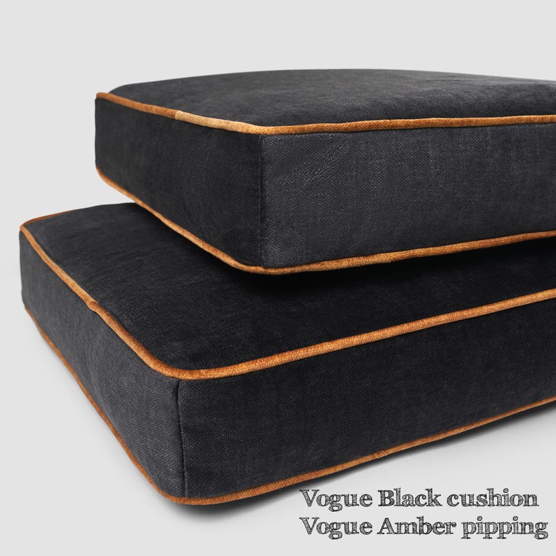 May include: Two black square cushions with amber-colored piping. The cushions are stacked on top of each other. The text "Vogue Black cushion Vogue Amber pipping" is at the bottom of the image.
