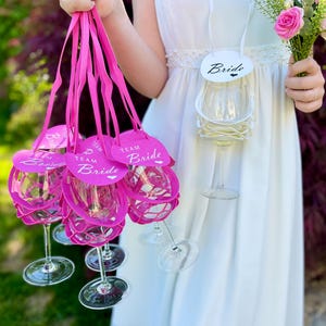 Könnte beinhalten: Klare Weingläser mit rosa Akzenten und Anhängern mit der Aufschrift "Team Bride". Ein Weinglas hat einen Anhänger mit der Aufschrift "Bride". Die Gläser werden von einer Person in einem weißen Kleid gehalten, die einen Blumenstrauß hält.