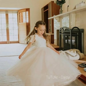 May include: A young girl in a light pink one-shoulder tulle dress is twirling. The dress has a detailed bodice and a flowing skirt. The text "TuTulle - Daisy" is visible. The girl has long blonde hair. The dress is a pale pink colour.
