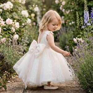 May include: A white sleeveless dress featuring a full tulle skirt with subtle sequin embellishments, a deep V-back, and a large decorative bow at the waist. The dress is paired with simple white flat shoes, creating a formal appearance.