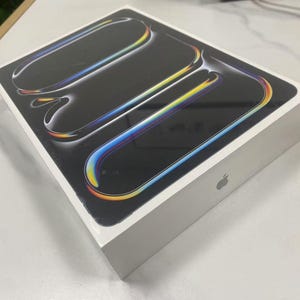 Puede incluir: Caja rectangular blanca con una parte superior negra que presenta tres diseños curvos, coloridos y reflectantes. La caja tiene el logotipo de Apple en el lateral. Los diseños tienen un degradado de colores, incluyendo amarillo, azul y naranja.