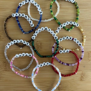 May include: A collection of beaded bracelets with white letter beads spelling out album titles like "Midnights," "Evermore," and "Reputation." The bracelets are in various colors, including blue, green, and red, and are arranged on a wooden surface.