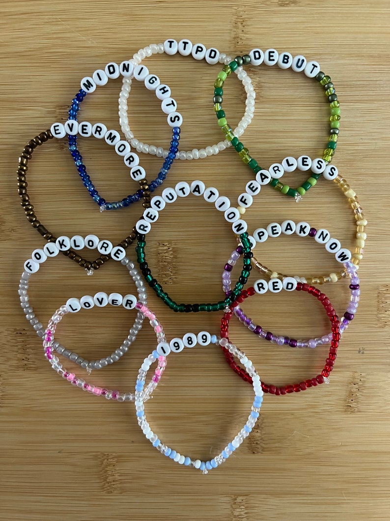 May include: A collection of beaded bracelets with white letter beads spelling out album titles like "Midnights," "Evermore," and "Reputation." The bracelets are in various colors, including blue, green, and red, and are arranged on a wooden surface.