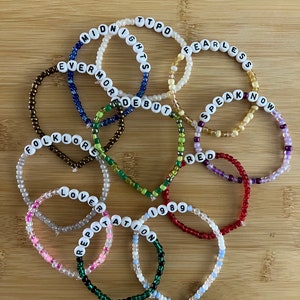May include: A collection of beaded bracelets with white letter beads spelling out song titles and album names. The bracelets are in various colors, including pink, green, blue, and red, and are arranged on a wooden surface.