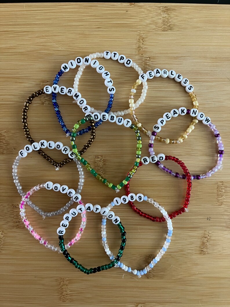 May include: A collection of beaded bracelets with white letter beads spelling out song titles and album names. The bracelets are in various colors, including pink, green, blue, and red, and are arranged on a wooden surface.