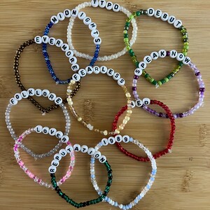 May include: A collection of beaded bracelets with white letter beads spelling out words and phrases like "MIDNIGHTS", "EVERMORE", and "FOLKLORE". The bracelets are in various colors, including white, blue, green, and red, and are arranged on a wooden surface.