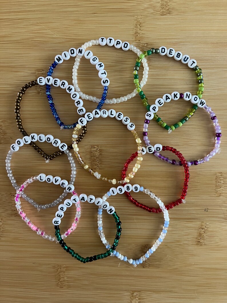 May include: A collection of beaded bracelets with white letter beads spelling out words and phrases like "MIDNIGHTS", "EVERMORE", and "FOLKLORE". The bracelets are in various colors, including white, blue, green, and red, and are arranged on a wooden surface.