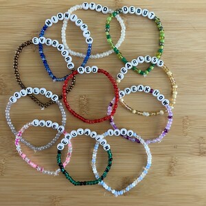 May include: A collection of beaded bracelets with white letter beads spelling out album titles like "Midnights," "Fearless," and "1989." The bracelets are in various colors, including red, green, and blue, and are arranged on a wooden surface.