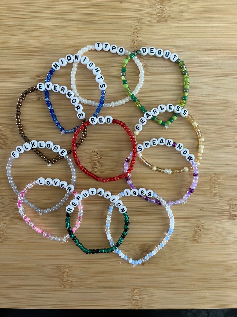 May include: A collection of beaded bracelets with white letter beads spelling out album titles like "Midnights," "Fearless," and "1989." The bracelets are in various colors, including red, green, and blue, and are arranged on a wooden surface.