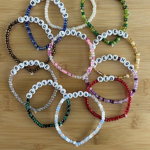 May include: A collection of beaded bracelets in various colors, each spelling out a word or phrase in white letter beads. The bracelets include the words "MIDNIGHTS", "EVERMORE", "FOLKLORE", "REPUTATION", "TTPD", "DEBUT", "FEARLESS", "SPEAK NOW", "RED", "1989", and "LOVER".