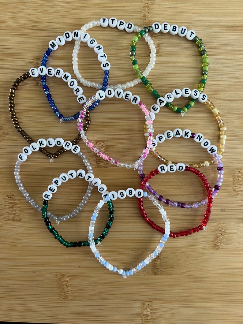 May include: A collection of beaded bracelets in various colors, each spelling out a word or phrase in white letter beads. The bracelets include the words "MIDNIGHTS", "EVERMORE", "FOLKLORE", "REPUTATION", "TTPD", "DEBUT", "FEARLESS", "SPEAK NOW", "RED", "1989", and "LOVER".