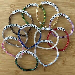 May include: A collection of beaded bracelets with white letter beads spelling out album titles like "Fearless," "Red," and "Reputation." The bracelets are in various colors, including red, green, and blue, and are arranged on a wooden surface.