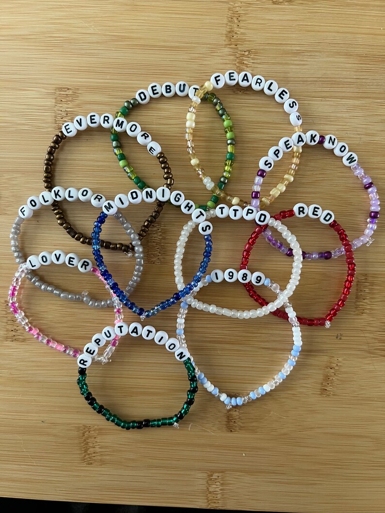 May include: A collection of beaded bracelets with white letter beads spelling out album titles like "Fearless," "Red," and "Reputation." The bracelets are in various colors, including red, green, and blue, and are arranged on a wooden surface.