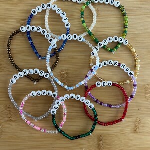 May include: A collection of beaded bracelets with white letter beads spelling out album titles like "Midnights," "Fearless," and "Reputation." The bracelets are in various colors, including green, red, and blue, and are arranged on a wooden surface.
