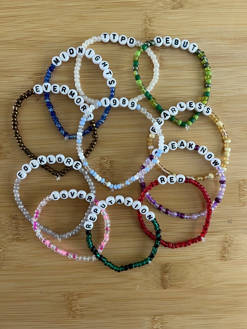May include: A collection of beaded bracelets with white letter beads spelling out album titles like "Midnights," "Fearless," and "Reputation." The bracelets are in various colors, including green, red, and blue, and are arranged on a wooden surface.