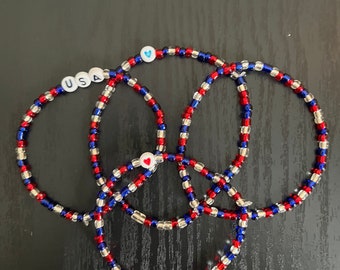 May include: Four beaded bracelets in red, white, and blue, with the word "USA" spelled out on one bracelet. Another bracelet has a heart-shaped bead, and another has a blue heart bead. The bracelets are arranged in a circular pattern.