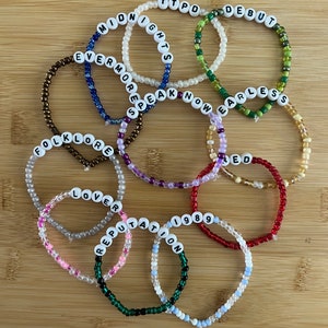 May include: A collection of beaded bracelets with white letter beads spelling out words like "Midnights", "Debut", "Evermore", "Folklore", "Lover", "Reputation", "1989", "Speak Now", "Fearless", "Red", and "TTPD". The bracelets are in various colors.