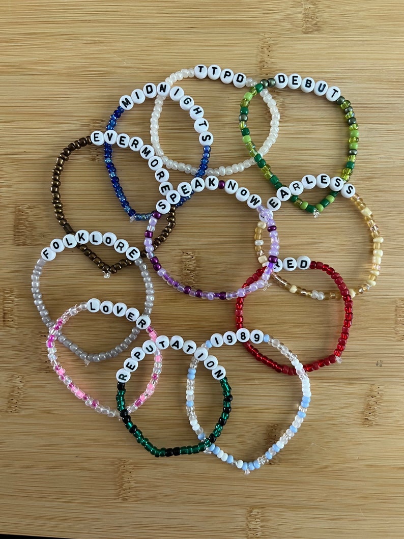 May include: A collection of beaded bracelets with white letter beads spelling out words like "Midnights", "Debut", "Evermore", "Folklore", "Lover", "Reputation", "1989", "Speak Now", "Fearless", "Red", and "TTPD". The bracelets are in various colors.
