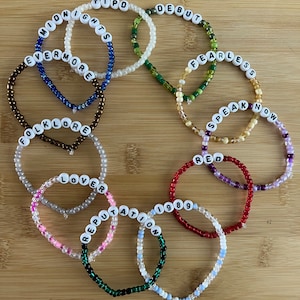 May include: A collection of beaded bracelets with white letter beads spelling out album titles like "Midnights," "Debut," and "Fearless." The bracelets feature various colors, including red, green, and pink, and are arranged on a wooden surface.