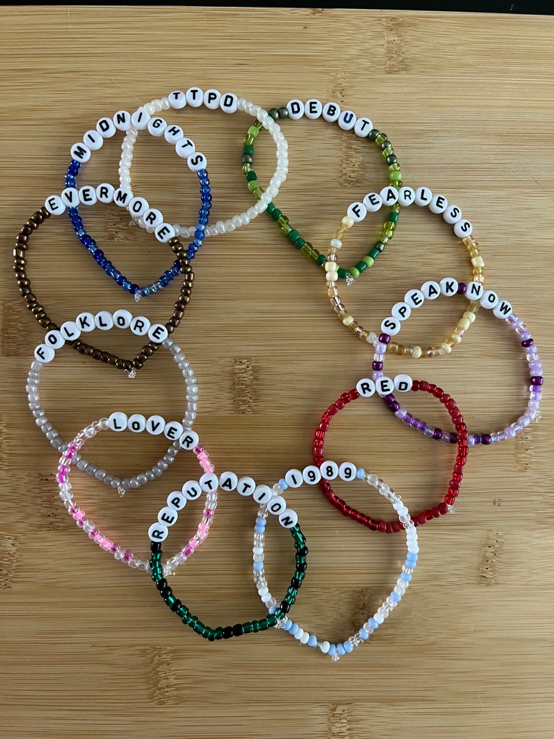 May include: A collection of beaded bracelets with white letter beads spelling out album titles like "Midnights," "Debut," and "Fearless." The bracelets feature various colors, including red, green, and pink, and are arranged on a wooden surface.