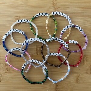 May include: A collection of beaded bracelets in various colors, each spelling out a different word. The bracelets are made with small, colorful beads and white letter beads. The words include "Debut", "Fearless", "Speak Now", "Red", and "1989".