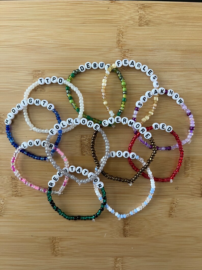 May include: A collection of beaded bracelets in various colors, each spelling out a different word. The bracelets are made with small, colorful beads and white letter beads. The words include "Debut", "Fearless", "Speak Now", "Red", and "1989".