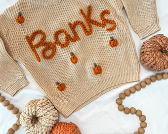 May include: A beige knitted sweater with the word "Thanks" embroidered in orange yarn. The sweater is decorated with small orange pumpkin appliques.