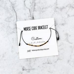 May include: A black Morse code bracelet with gold and black beads is displayed on a white card. The card reads "MORSE CODE BRACELET" and "Custom". The Etsy shop URL is visible. The bracelet is on a marble surface.