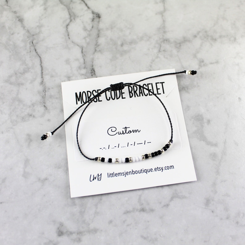 May include: Black and white beaded bracelet on a black string with a card that says "Morse Code Bracelet" and "Custom" with the morse code for "Custom" written below. The card also has the text "My littlemsjenboutique.etsy.com".
