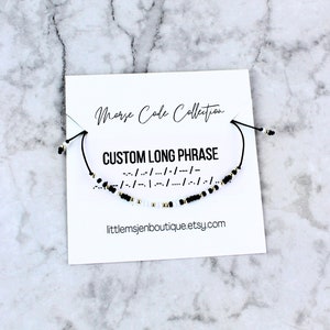 May include: A black and white beaded bracelet with a black cord. The bracelet is on a white card with the text "Morse Code Collection" and "CUSTOM LONG PHRASE" in black text. The card also includes the morse code for the phrase and the website "littlemsjenboutique.etsy.com".