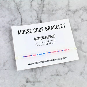 May include: A white card with the text "MORSE CODE BRACELET" and "CUSTOM PHRASE" printed on it. The card also includes a morse code message and a website address: "www.littlemsjenboutique.etsy.com". A white beaded bracelet with pink and blue beads is displayed on the card.