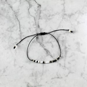 May include: A black and white beaded bracelet with a black cord and adjustable sliding knot closure.