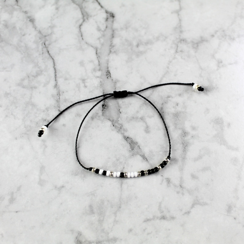 May include: A black and white beaded bracelet with a black cord and adjustable sliding knot closure.