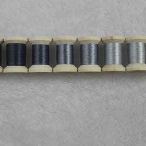 May include: A row of seven wooden spools, each holding a different shade of gray thread. The thread colors range from dark charcoal to light silver. The spools are arranged horizontally against a soft, light gray background.