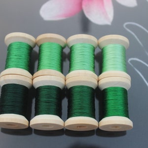 May include: Eight wooden spools of green thread are stacked in pairs. The thread colors range from dark forest green to a lighter, brighter green. The spools have a natural wood finish.