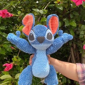 May include: A handmade blue crochet plush toy with large black eyes, a black nose, and large orange ears. The plush toy is in a standing position with its arms outstretched. The background features green foliage and pink flowers.