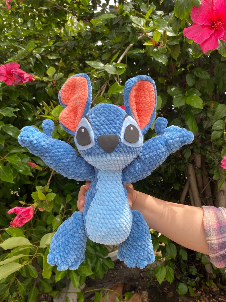 May include: A handmade blue crochet plush toy with large black eyes, a black nose, and large orange ears. The plush toy is in a standing position with its arms outstretched. The background features green foliage and pink flowers.