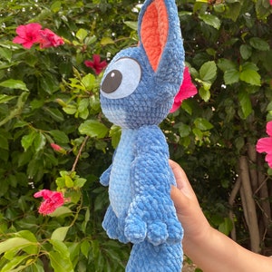 May include: A blue crochet plush toy with large ears and a big eye. The plush toy has a light blue body and darker blue accents. The inside of the ears is orange. The toy is held in front of a background of green leaves and pink flowers.