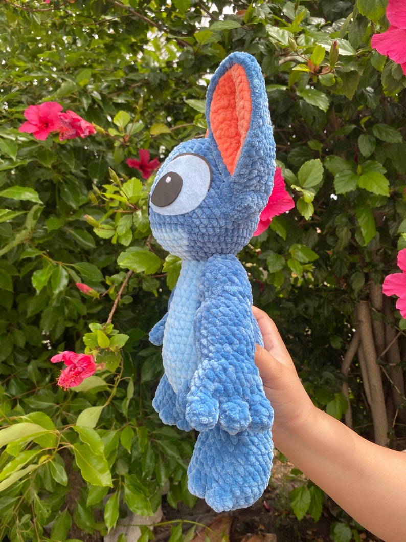 May include: A blue crochet plush toy with large ears and a big eye. The plush toy has a light blue body and darker blue accents. The inside of the ears is orange. The toy is held in front of a background of green leaves and pink flowers.