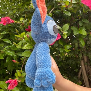 May include: A blue plush toy with large ears and a distinctive eye design. The toy has a textured, fuzzy appearance and is held against a backdrop of green foliage and pink flowers. The toy's ears are lined with a peach-colored fabric.