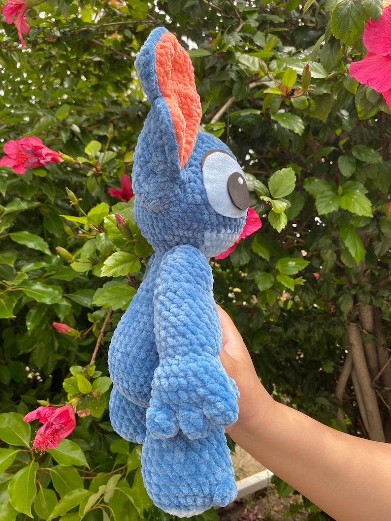 May include: A blue plush toy with large ears and a distinctive eye design. The toy has a textured, fuzzy appearance and is held against a backdrop of green foliage and pink flowers. The toy's ears are lined with a peach-colored fabric.