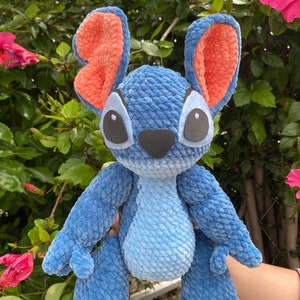 May include: A handmade blue crochet plush toy with large ears, featuring orange inner ears, and black eyes. The plush has a light blue belly and a black nose. The toy is held in front of a green leafy background with pink flowers.